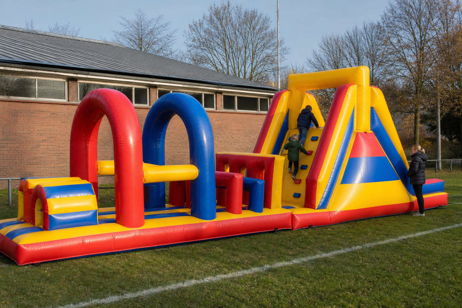 De Stormbaan ligt als een lang rood-geel-blauw parcours op een schoolveld in Limburg, met kinderen die van achteren de klimmuur op gaan.